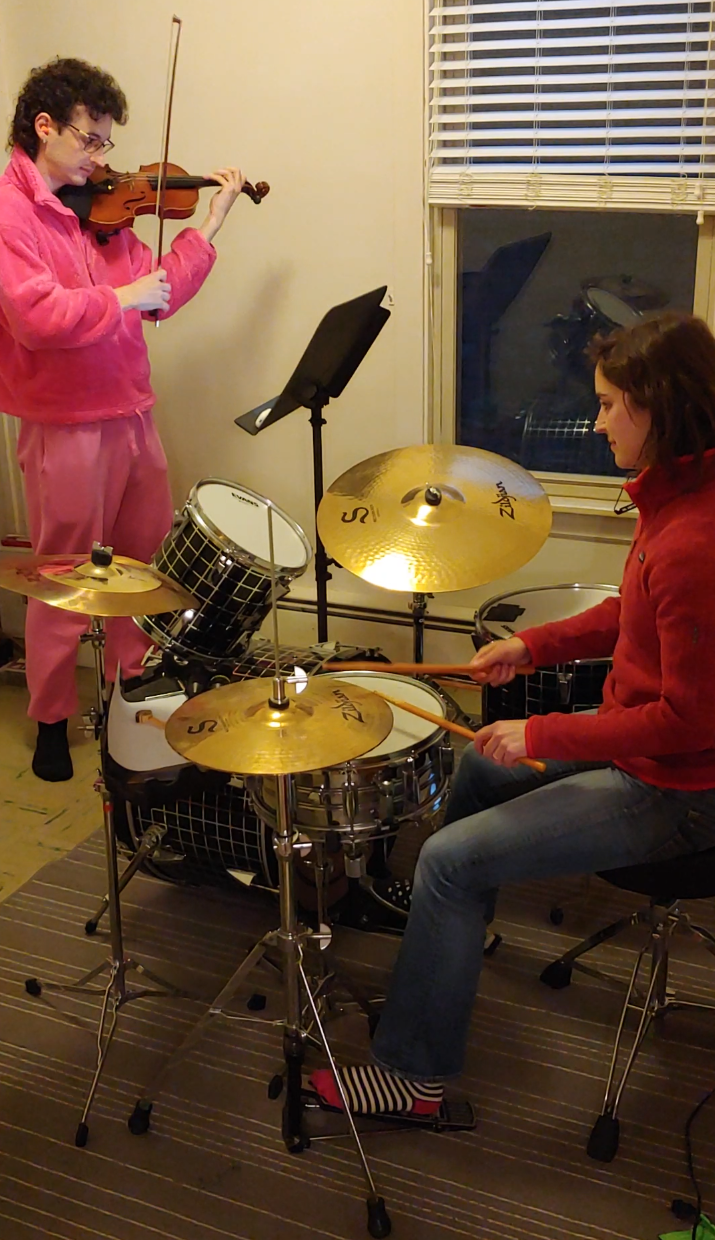  A tall, dark-haired person stands playing violin. A brown-haired woman sits at a drum set, playing the drums. They are in a spare room in a house. 