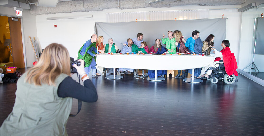 Artists gather behind a table to recreate the Last Supper painting. A photographer is framing up the picture.